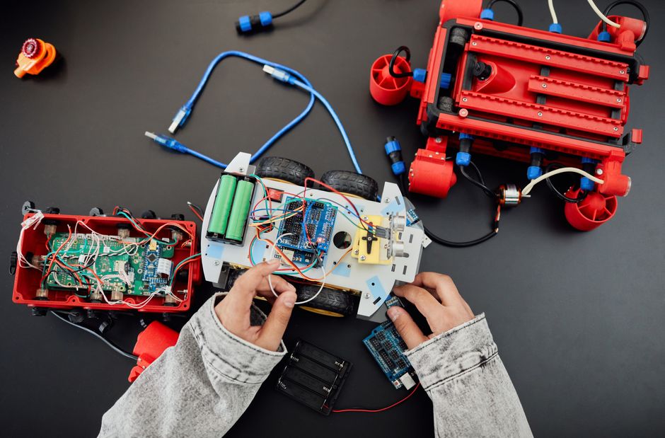 A student's hands wiring an Arduino-powered robot cart on a dark lab bench, two finished bots beside it